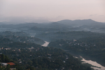 Aerial view of the Mahaweli River flowing through a valley surrounded by lush green hills and the city of Kandy, Sri Lanka, during a misty sunrise