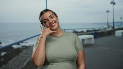Young plus-size woman making call me gesture at seaside with beach and ocean in background,...