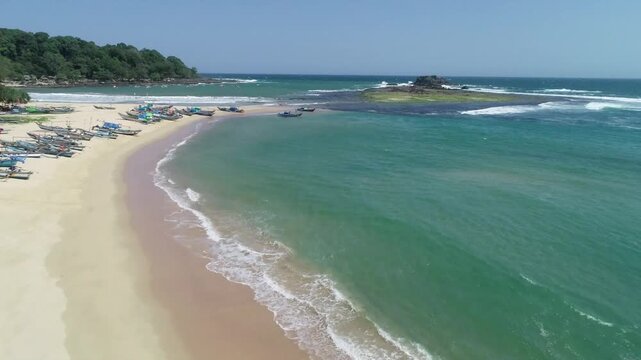 Drone aerial view of a tropical beach with golden sand, turquoise sea, fishing boats, and small island under clear blue sky.