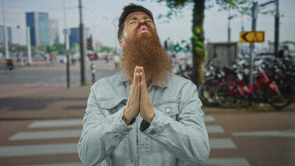 Bearded man with clasped hands in blue denim jacket gazes skyward beside bike rack on busy city...