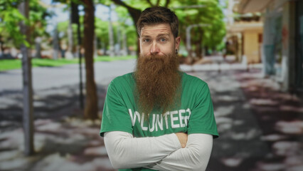 Man with long brown beard wearing green volunteer shirt and arms crossed on sunny street lined with...