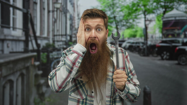Man holding wrench with one hand and raising other hand on street, wearing plaid shirt and sporting a long beard; surprise. - Powered by Adobe