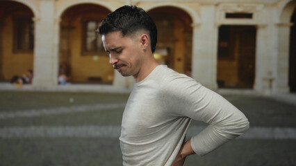 Obraz premium Hispanic man in pain holds his back outdoors in an old university setting, wearing casual attire with arches in the background, conveying discomfort and strain.