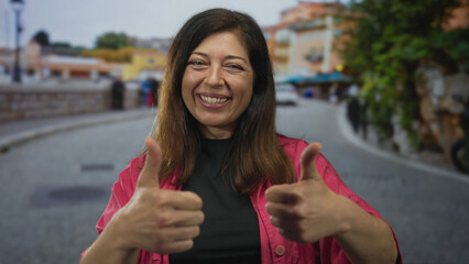 Middle-aged woman in pink jacket shows dual thumbs up gesture on narrow cobblestone street in city...