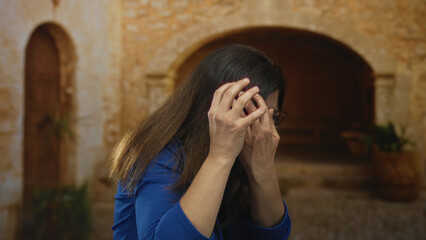 Woman in blue top covers face with both hands under stone archway on old town street corner;...