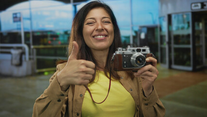 Middle age hispanic woman holds vintage camera and shows thumbs up at airport terminal outdoors;...