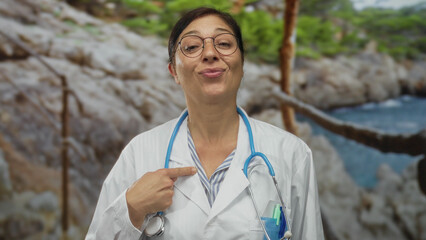 Woman doctor in white coat points finger to chest with stethoscope at sunny seaside beach;...