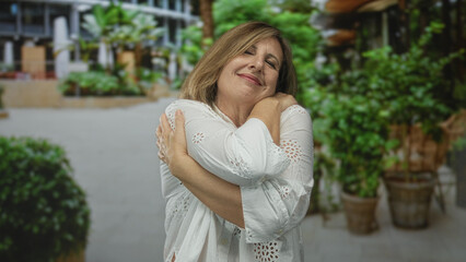 Mature woman smiling and hugging her arms on a tree lined street in front of potted plants; self...