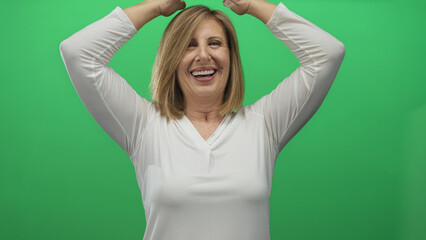 Middle aged woman wearing white blouse raises hands above head in studio with bright green...