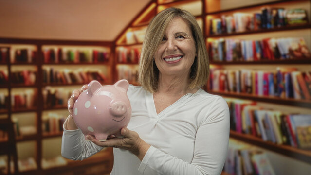 Middle aged woman holds pink piggybank with bare hands in building lined with bookshelves; financial security contentment.