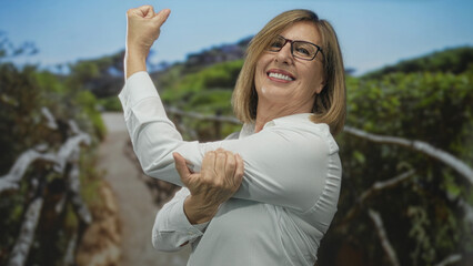 Woman wearing glasses pulls elbow in green forest under sunlight while smiling; vitality.