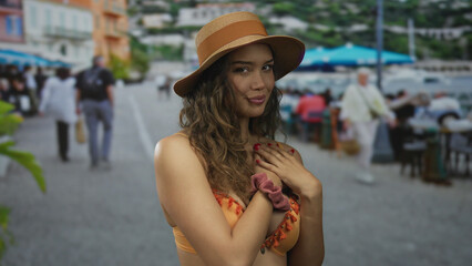 Young hispanic woman in sunhat and orange bikini makes finger heart gesture on street outside seaside restaurant; affection.