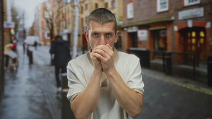 Man feeling cold on a wet street with hands clasped, wearing a light shirt, against a blurred city...