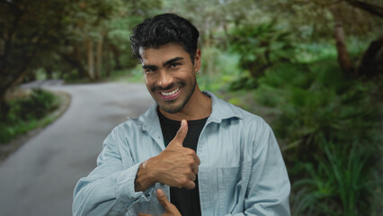 Man shows thumb up gesture on a winding sunlit forest path lined with lush trees while smiling;...