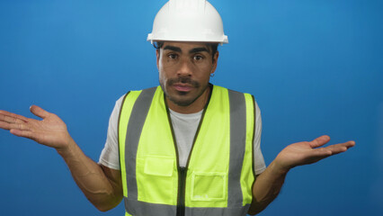 Man with safety vest and hardhat shrugs palms up against solid blue backdrop in modern studio;...
