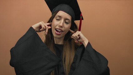 Young hispanic woman wearing black graduation gown and mortarboard covers ears with fingers in...