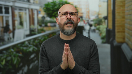 Caucasian man with glasses and beard gestures pleadingly in an urban outdoor setting with blurred...