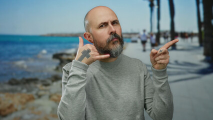 Bald bearded man gesturing the phone symbol on a seaside promenade with the ocean in the background...