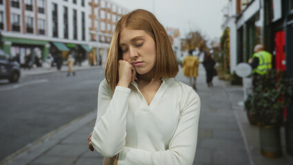 Fototapeta premium Woman standing on an urban street with closed eyes, expressing calm and thoughtfulness, against a backdrop of city life and modern buildings, during a slightly overcast day.