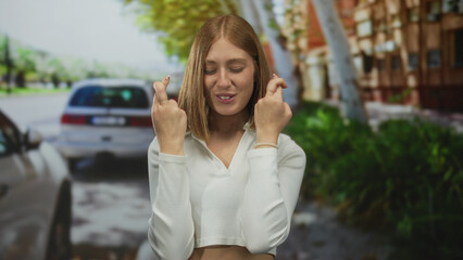 Fototapeta premium Woman outdoors crossing fingers in hope on an urban street with parked cars and trees in the background, reflecting a positive and hopeful expression in a city environment.