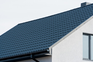 Side view of a house shows the dark blue metal roof, a black gutter, white stucco walls, and a...