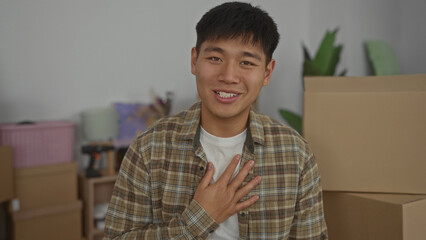 Obraz premium Young man smiling in new home living room surrounded by moving boxes expressing excitement and positivity in a cozy indoor apartment setting.