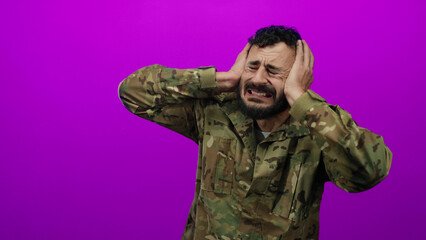 Hispanic man in military uniform holds head in distress against vivid pink background, conveying emotion and intensity.