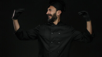 Hispanic male chef with a beard gestures expressively wearing black gloves and uniform against a dark background, conveying culinary expertise and passion.
