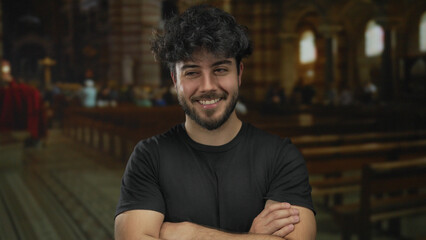 Young hispanic man with beard in casual clothes stands smiling with arms crossed inside a dimly lit...