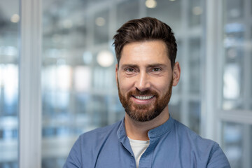 Obraz premium A cheerful bearded man wearing a blue shirt is seen in a bright office setting, radiating positivity and professional confidence with a welcoming demeanor.