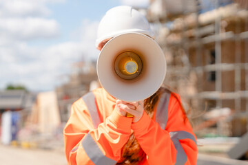 Portrait of young woman construction worker in vivid safety gear uses a megaphone to communicate...