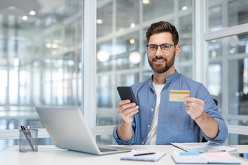 Business professional holding a smartphone and a credit card, working at a bright office desk.