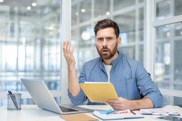 A professional man in a modern office, reacting with disbelief while reviewing a folder of...