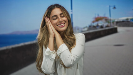 Young blonde woman holds both hands to her cheek in a resting gesture on seaside promenade; rest...