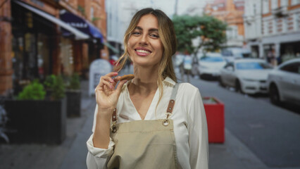 Young blonde woman wearing apron holds cookie between index and thumb outside cafe on city street;...
