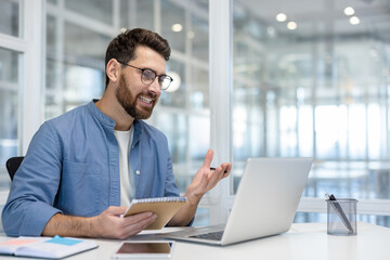 A smiling man in glasses holds a notebook during a video conference in a modern office space, showcasing communication, professionalism, and a tech-driven work environment.