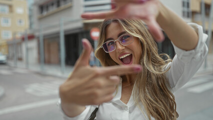 Young blonde woman smiles and makes finger frame to camera in urban street; creativity perspective...