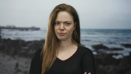 Woman with arms crossed and closed eyes standing beside rocky seaside shore under overcast sky;...