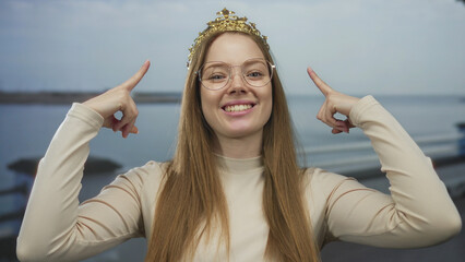 Young woman wearing golden crown points fingers to crown on street by seaside railing; joy...