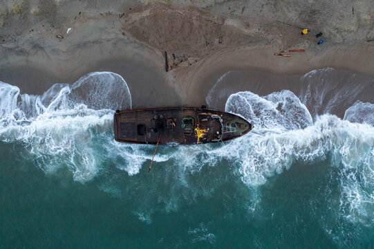 Aerial view of a rusting ship stranded on the sandy shore, waves crashing against its hull, a stark contrast of decay against the vibrant sea, Anapa, Krasnodar Krai, Russia.