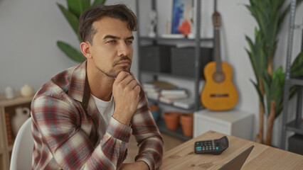 Young man in an office setting analyzing data at a wooden desk with a laptop and guitar in the background conveying focus and contemplation indoors