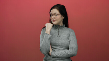 Brunette woman in glasses and grey sweater confidently gestures with thumb against vibrant red background, embodying beauty and assertive expression.