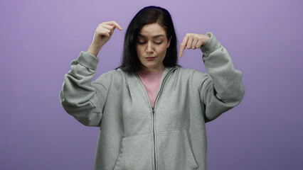 Fototapeta premium Woman with brunette hair wearing gray sweatshirt posing against a purple background expressing pointing gestures and looking curious.