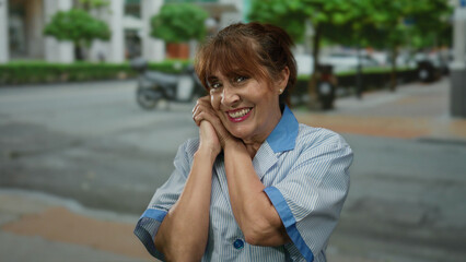 Smiling senior hispanic woman wearing cleaner uniform on a city street with buildings and trees in...
