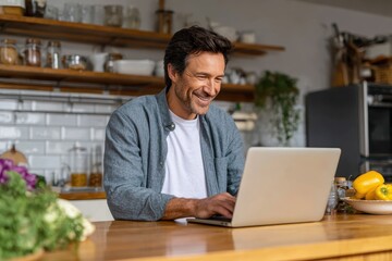 Fototapeta premium Working From Kitchen Table. Young Man Smiling and Working on Laptop Computer at Home