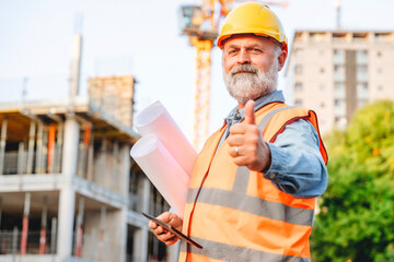 A construction site supervisor stands confidently at a development site, holding blueprints and...