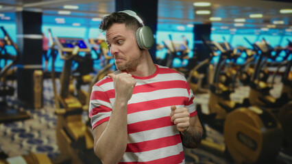 Young man with headphones making a fist in a gym with exercise machines blurred in the background...