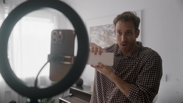 Social media influencer and content creator reviewing product or unboxing video. Talking to smart phone filming him with ring light stand in apartment or home studio. Excited and happy filming video