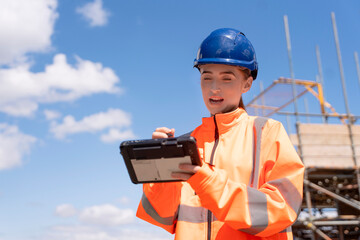 Portrait of smiling young woman construction worker in bright safety gear using rugged tablet at...
