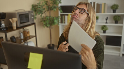Man holds white document to forehead at desk in cluttered office with computer monitor and shelves;...